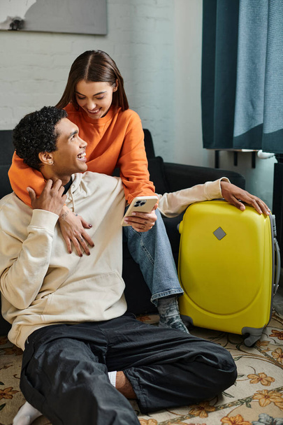 happy woman holding smartphone and hugging african american boyfriend near luggage in hostel room - Photo, Image