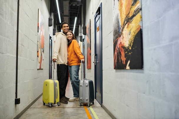 Coppia multiculturale rilassata con bagagli sorridente e in piedi insieme in un corridoio ostello - Foto, immagini