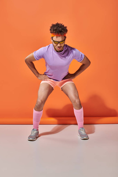 sportovní africký Američan muž v sluneční brýle a tělocvična oblečení cvičení na oranžovém pozadí - Fotografie, Obrázek