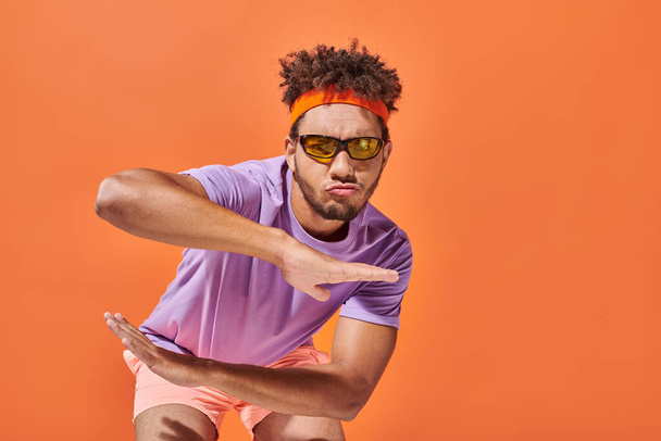 deportista afroamericano confiado en traje de gimnasio y gafas de sol gestos sobre fondo naranja - Foto, imagen
