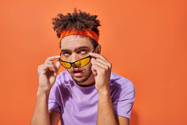 Gözlüklü ve bantlı gözlüklü kızgın Afrikalı Amerikalı adam turuncu arka planda yuvarlanan gözler, duygu - Fotoğraf, Görsel