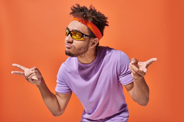 Gözlüklü, bantlı, dudak büken ve turuncu arka planda el kol hareketi yapan Afro-Amerikan bir adam. - Fotoğraf, Görsel