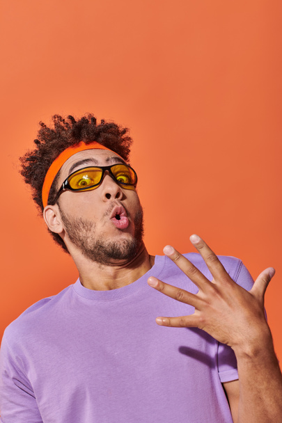 překvapený africký Američan muž v brýlích a čelenka při pohledu na kameru na oranžovém pozadí - Fotografie, Obrázek