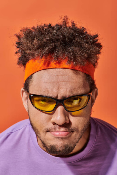 mladý afroameričan v brýlích a čelence při pohledu na kameru na oranžovém pozadí, zamračený - Fotografie, Obrázek