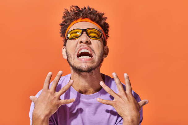 emotioneel afrikaanse man in hoofdband en zonnebril gebaren op oranje achtergrond - Foto, afbeelding