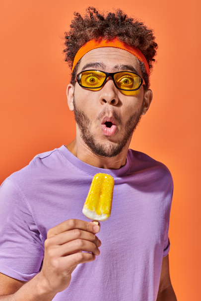 schockierter afrikanisch-amerikanischer Mann mit Sonnenbrille und Stirnband mit fruchtigem Eis auf orangefarbenem Hintergrund - Foto, Bild