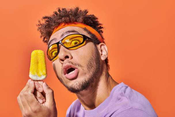 chocado homem americano africano em óculos de sol segurando sorvete congelado no fundo laranja - Foto, Imagem