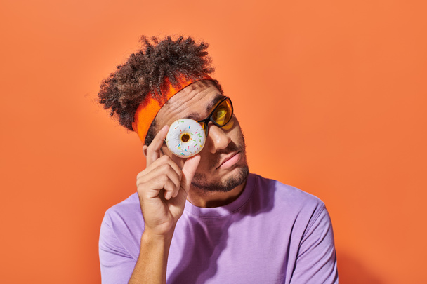 junger afrikanisch-amerikanischer Mann mit Sonnenbrille blickt durch ein Donut-Loch auf orangefarbenem Hintergrund in die Kamera - Foto, Bild