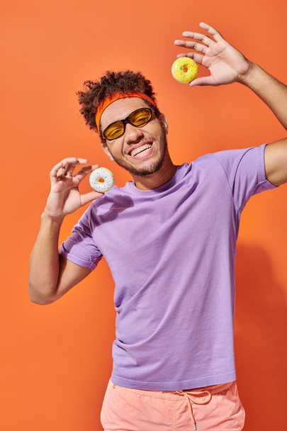 feliz joven afroamericano hombre en gafas de sol sosteniendo donuts tamaño mordida sobre fondo naranja - Foto, imagen