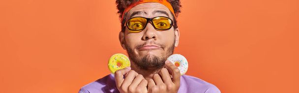 Güneş gözlüklü komik Afro-Amerikan adam turuncu arka planda ısırık büyüklüğünde çörekler tutuyor. - Fotoğraf, Görsel