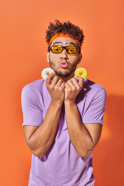 jong Afrikaans amerikaans man in zonnebril houden beet-sized donuts op oranje achtergrond, grimas - Foto, afbeelding