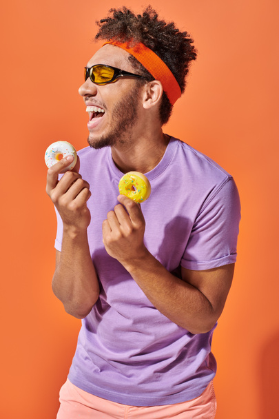 jong Afrikaans amerikaans man in zonnebril houden beet-sized donuts en lachen op oranje achtergrond - Foto, afbeelding
