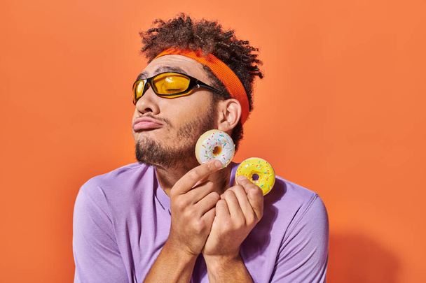 african american man in sunglasses holding bite-sized donuts on orange background, grimace - Photo, Image