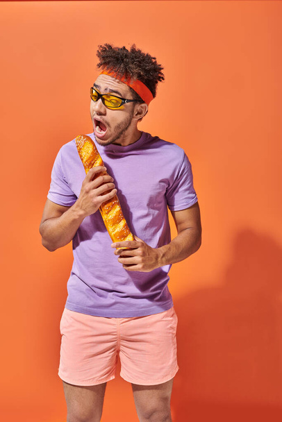 hombre americano africano divertido en gafas de sol y diadema mordiendo baguette fresca sobre fondo naranja - Foto, imagen