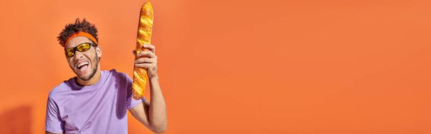 hombre afroamericano feliz en gafas de sol sosteniendo baguette fresca sobre fondo naranja, bandera - Foto, Imagen