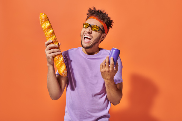 happy african american man in sunglasses holding fresh baguette and soda on orange background - Foto, afbeelding