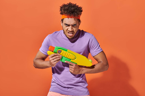 african american man in headband playing water fight with toy gun on orange background, grimace - Photo, Image