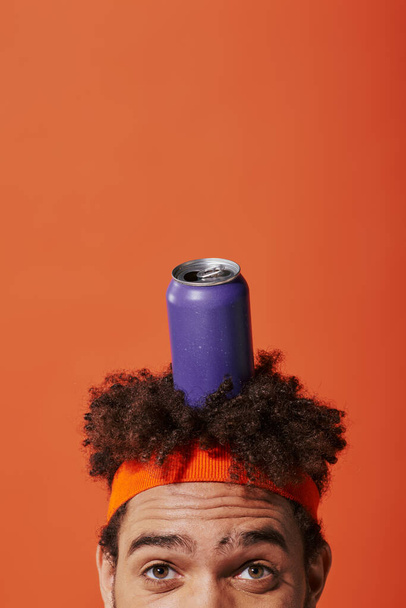 lila Getränkedose auf dem Kopf des lockigen afrikanisch-amerikanischen Mannes mit Stirnband auf orangefarbenem Hintergrund - Foto, Bild