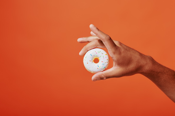 cropped shot of person holding glazed vanilla donut with sprinkles on orange background, white icing - Photo, Image
