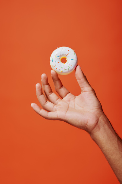 tiro recortado de hombre sosteniendo donut de vainilla vidriada con aspersiones sobre fondo naranja, glaseado blanco - Foto, imagen
