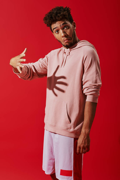 verwarde Afro-Amerikaanse man in hoodie gebaren en kijken naar camera op rode achtergrond, schaduw - Foto, afbeelding