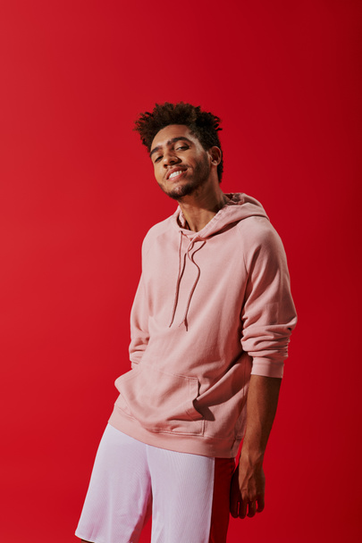 happy african american man in pink hoodie looking at camera while smiling on red background - Photo, Image