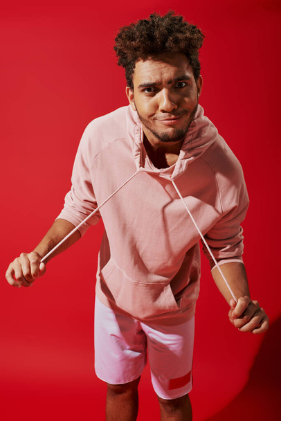 funny african american man pulling drawstrings of hoodie and looking at camera on red background - Photo, Image