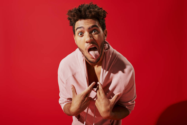 funny african american man pulling pink hoodie and sticking out tongue while on red background - Photo, Image