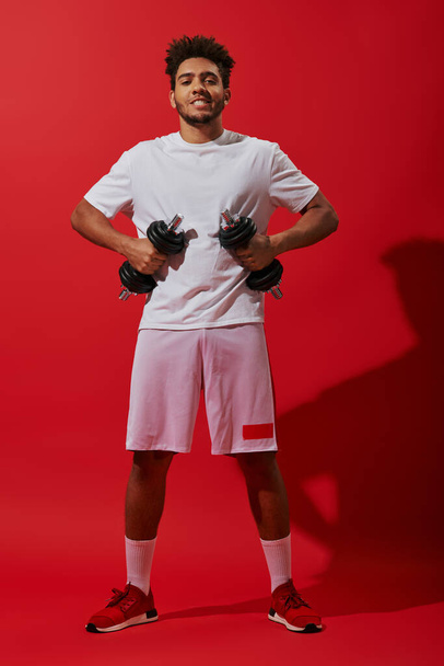 volle Länge des afrikanisch-amerikanischen Sportlers, der mit Kurzhanteln auf rotem Hintergrund trainiert, kraftvoll - Foto, Bild