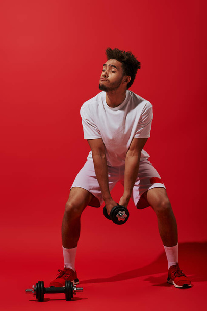 longitud completa de divertido afroamericano deportista ejercicio con mancuerna sobre fondo rojo - Foto, Imagen