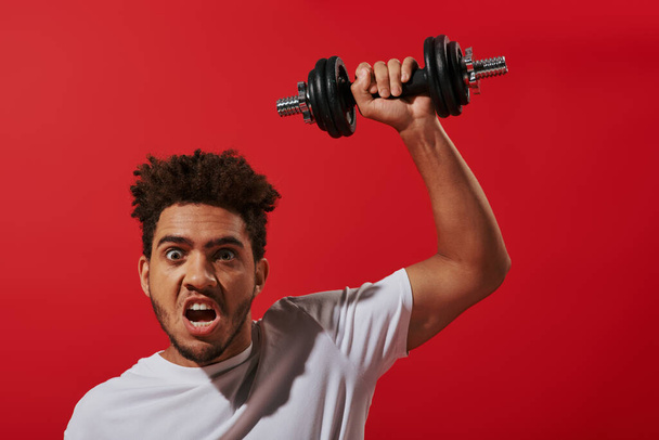 motivated african american man in sportswear working out with heavy dumbbell on red background - Fotografie, Obrázek