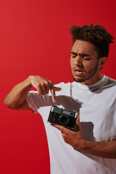 joven africano americano hombre mirando su vintage cámara en rojo fondo, pulsando botón - Foto, imagen