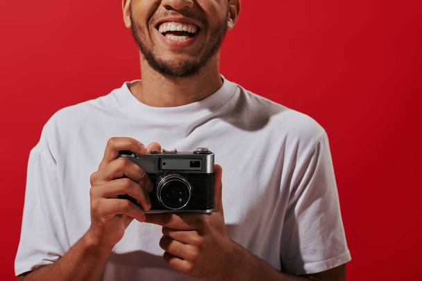 обрізаний і щасливий афроамериканський фотограф тримає старовинну камеру і посміхається на червоному тлі - Фото, зображення