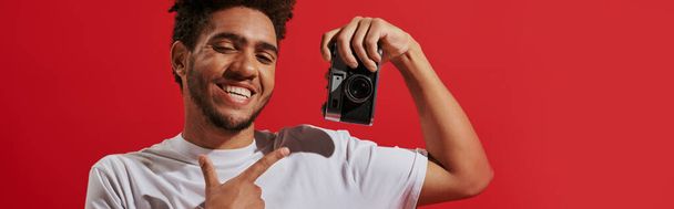 レトロカメラを指し,赤い背景に微笑んでいる幸せなアフリカ系アメリカ人の写真家,バナー - 写真・画像