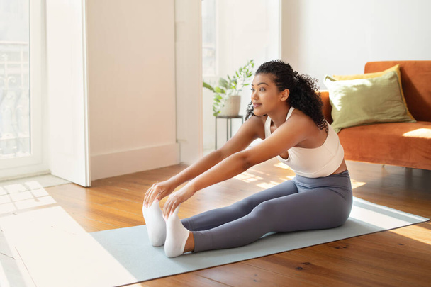 Fit young woman in sportswear stretching on yoga mat, performing seated toe touch exercises in her living room, embodying active and healthy lifestyle in morning fitness routine - Photo, image