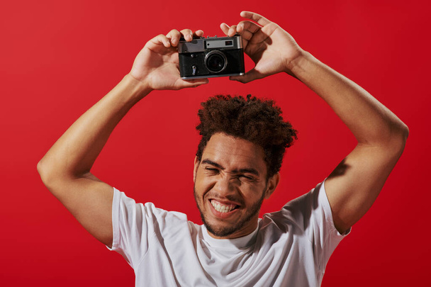 vreugdevolle Afrikaanse amerikaanse fotograaf met retro camera en glimlach op rode achtergrond - Foto, afbeelding