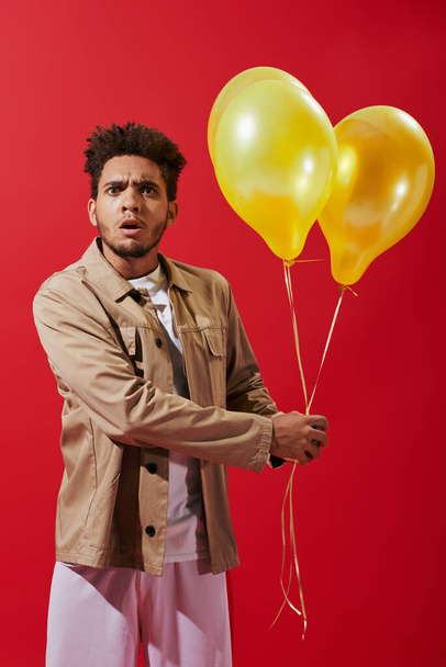geschokt Afrikaan amerikaanse man in beige jas houden helium ballonnen op rode achtergrond, partij - Foto, afbeelding