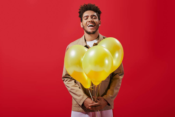 veselý africký Američan v béžové bundě drží balónky a směje se na červeném pozadí - Fotografie, Obrázek