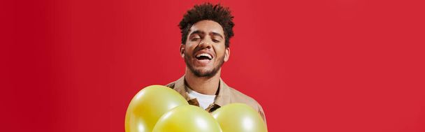 felice uomo afroamericano in giacca beige che tiene palloncini e ride su sfondo rosso, striscione - Foto, immagini