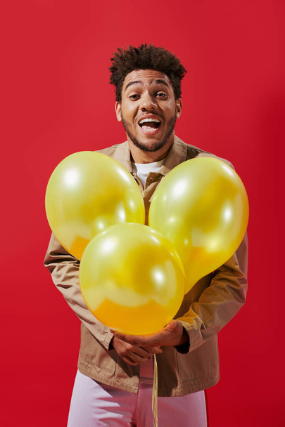 positieve Afro-Amerikaanse man in beige jas houden ballonnen en lachen op rode achtergrond - Foto, afbeelding