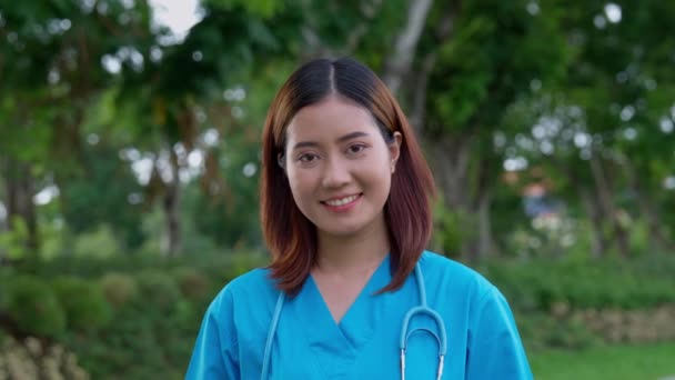Retrato de mujer médica asiática confiada, feliz y sonriente médico o enfermera con uniforme de uniformes azules, al aire libre - Imágenes, Vídeo