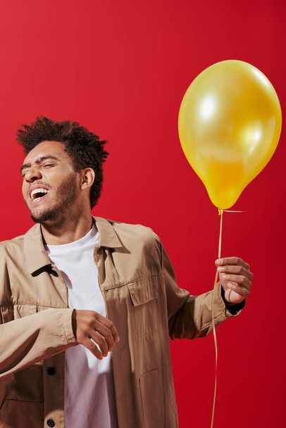 optimistic african american man in beige jacket holding balloon and smiling on red background - Photo, Image
