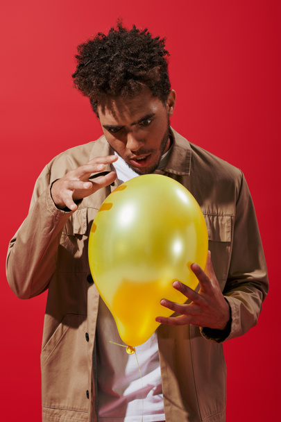 enchanted african american man in beige jacket looking at balloon on red background, amusement - Photo, Image