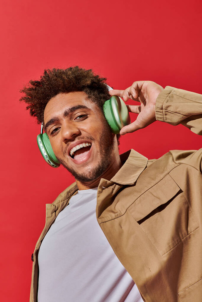 portrait of excited african american fella in wireless headphones listening music on red background - Photo, Image