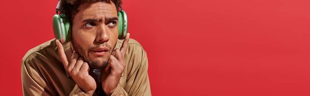 bannière de l'homme afro-américain confus dans les écouteurs sans fil écouter de la musique sur fond rouge - Photo, image