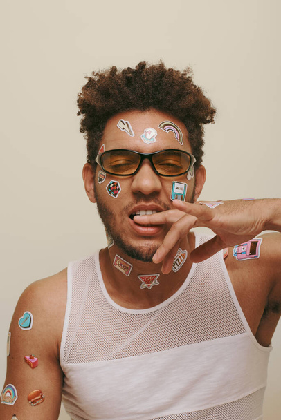young african american man in sunglasses with trendy stickers on face biting finger on grey backdrop - Photo, Image