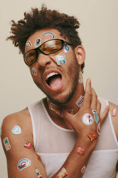 expressieve Afrikaans-Amerikaanse man in zonnebril met trendy stickers op gezicht op grijze achtergrond - Foto, afbeelding