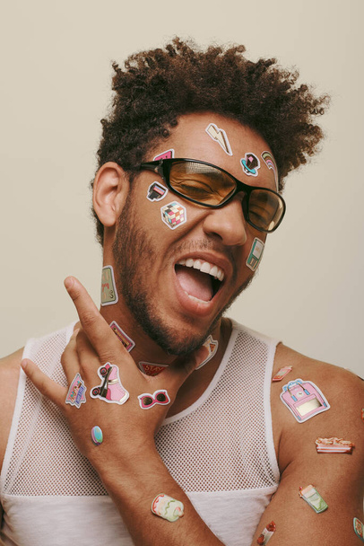 expressieve Afrikaans Amerikaanse kerel in zonnebril met trendy stickers op gezicht op grijze achtergrond - Foto, afbeelding