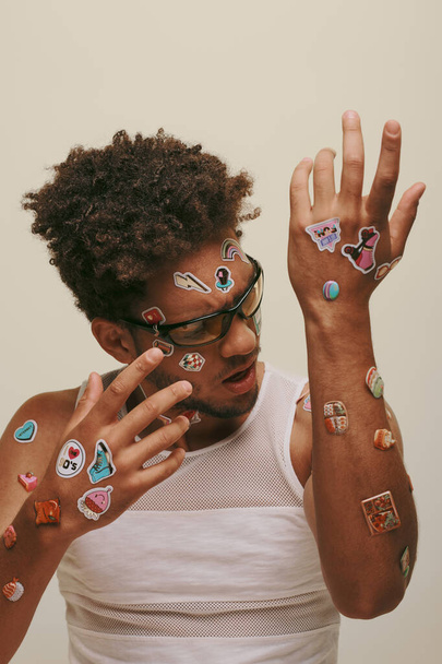 verbaasd Afrikaans amerikaanse man in een zonnebril kijken naar trendy stickers op zijn lichaam op grijze achtergrond - Foto, afbeelding
