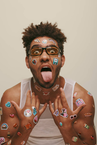 excentrieke jonge Afrikaans-Amerikaanse man in zonnebril met trendy stickers op gezicht op grijze achtergrond - Foto, afbeelding